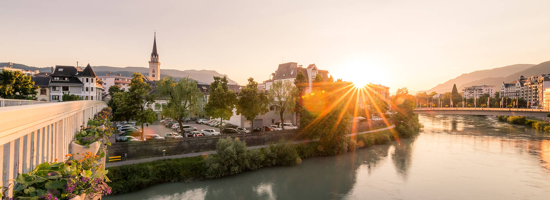 Das Bild zeigt einen stimmungsvollen Blick auf Villach, die zweitgrößte Stadt Kärntens, während eines Sonnenuntergangs an der Drau.