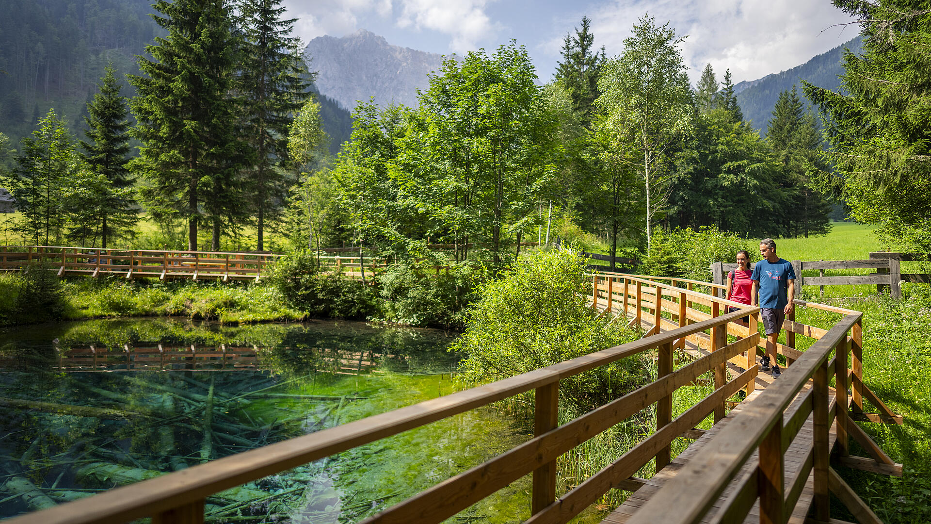 Zwei Wanderer spazieren auf einem Holzsteg entlang des türkisfarbenen Meerauges im Bodental, umgeben von grüner Natur, Bäumen und Bergkulisse.